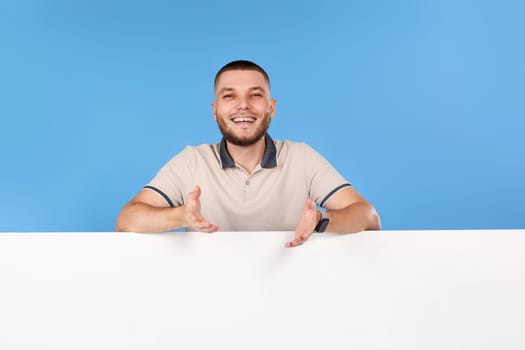 Portrait of cheerful bearded man leaning on empty placard for advertisement with empty copy space on blue background