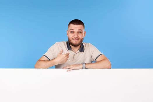 man pointing at himself and shrugging confused with board with empty copy space isolated on blue background
