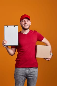 friendly delivery man in red cap, t-shirt holding clipboard and empty cardboard box isolated on orange background
