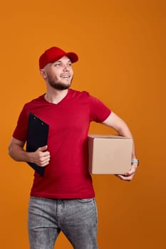 Delivery man in red cap, t-shirt holding clipboard and empty cardboard box isolated on orange background