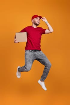 Delivery man in red cap, t-shirt holding cardboard box and jumping isolated on orange background