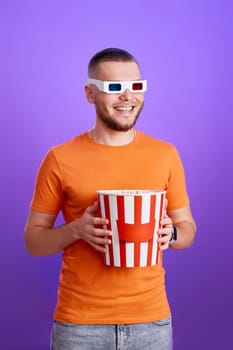 Young handsome caucasian man in 3d glasses holding a big bucket of popcorns on purple background