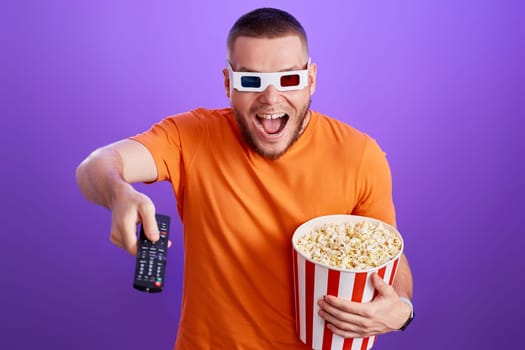 surprised man in 3d glasses using remote controller watching movie and holding big bucket of popcorns on purple background
