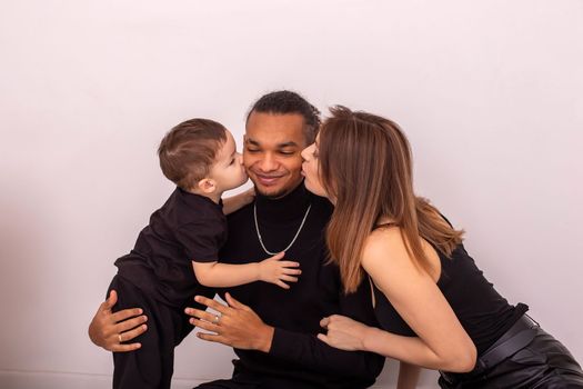 a happy family, a woman and a little boy, in black clothes, kiss a stylish young man on the cheeks from both sides, sit near a light wall. Copy space. Close up