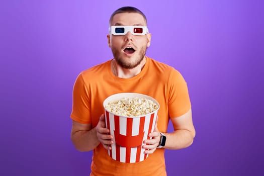 amazed caucasian man in 3d glasses holding a big bucket of popcorns on purple background