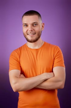 smiling young guy in orange t-shirt posing with crossed arms on purple studio background