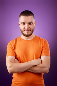 smiling young guy in orange t-shirt posing with crossed arms and looking at the camera on purple studio background