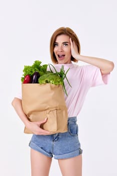 Young surprised blonde vegetarian woman in casual clothes hold paper bag with vegetables on white background