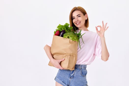 cheerful happy blonde woman hold paper bag with vegetables and showing Ok gesture on white background. copy space
