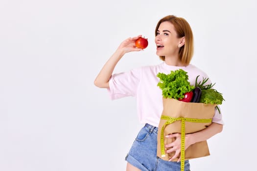 beautiful blonde woman in pink t-shirt hold paper bag with vegetables and red apple on white background. copy space