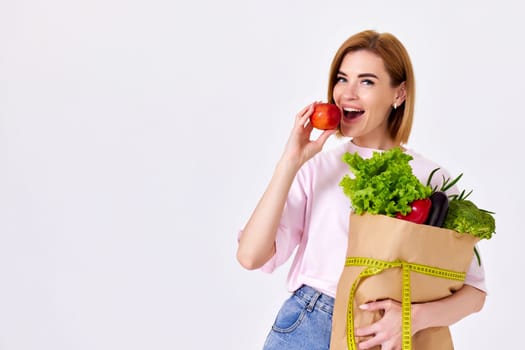 smiling caucasian woman in pink t-shirt with paper bag with vegetables biting red apple. copy space. healthy food for diet