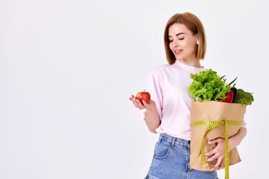 young caucasian woman in pink t-shirt hold paper bag with groceries and with measuring tape and red apple on white background. copy space