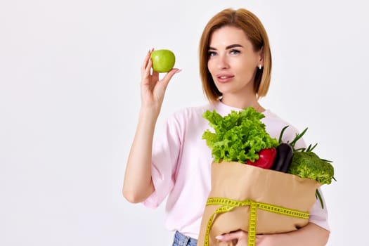 young caucasian woman in pink t-shirt holds paper bag with vegetables and shows green apple on white background. copy space