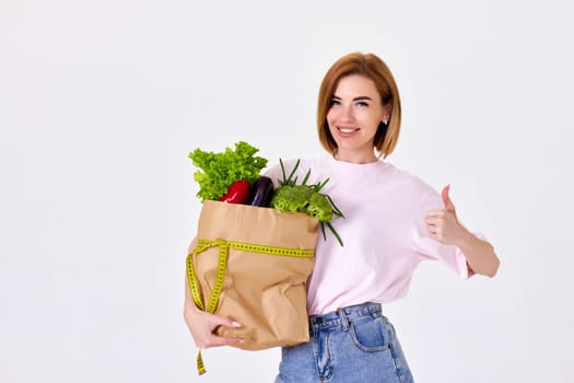 smiling caucasian sporty woman holds paper bag with vegetables, measuring tape and showing Ok gesture on white background. copy space