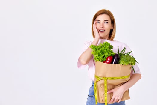 surprised slim woman holds paper bag with vegetables and measuring tape on white background. copy space. healthy food