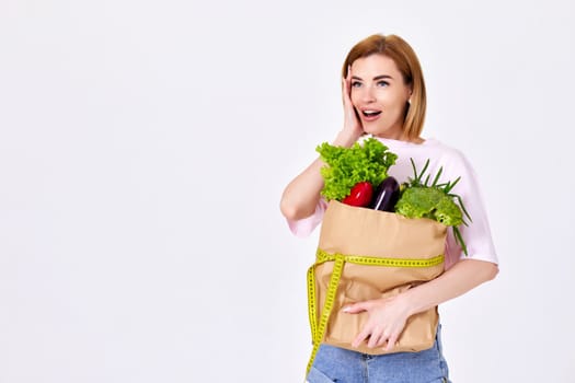 surprised slim woman hold paper bag with vegetables and measuring tape on white background. copy space. Beautiful girl eating healthy food for diet