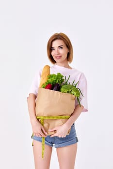 sporty slim woman holding a shopping bag full of groceries and measuring tape on white background.