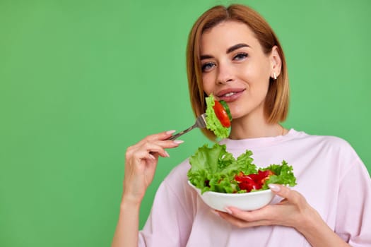 beautiful smiling blonde woman eating fresh vegetable salad on green background. copy space