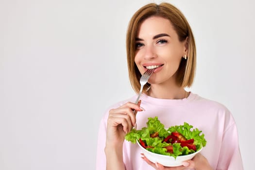 smiling healthy blonde woman eating fresh vegetable salad on white background. copy space