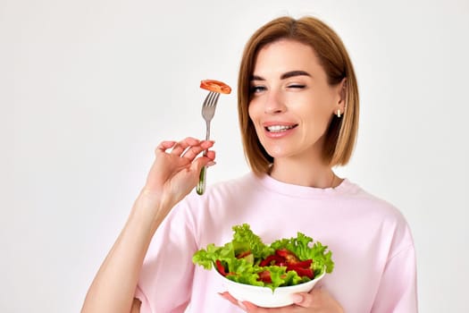 beautiful smiling blonde girl eating fresh vegetable salad and winking on white background