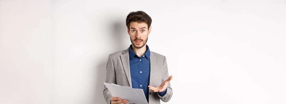 Confused businessman looking at bad document, complaining on deal, pointing at paper frustrated, standing in suit on white background.