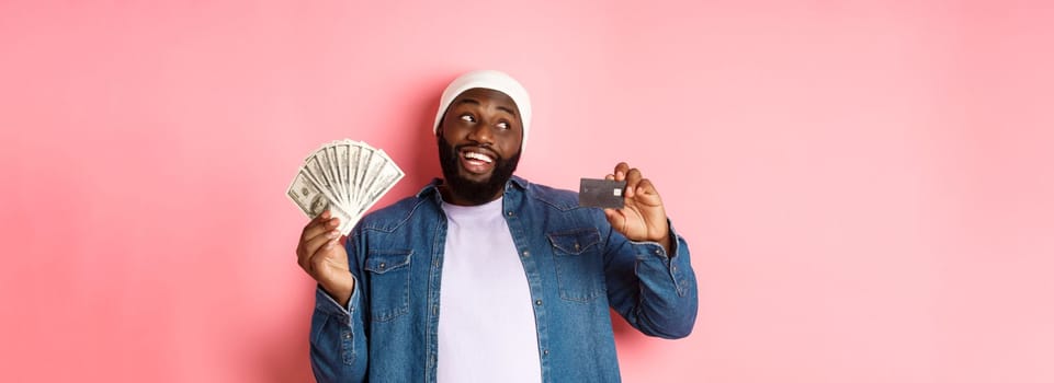 Dreamy african-american man showing credit card and dollars, thinking about shopping and smiling, standing over pink background.