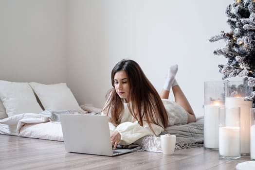 Merry Christmas and Happy New Year. Woman in warm white winter sweater lying in bed at home using laptop, watching movies or working