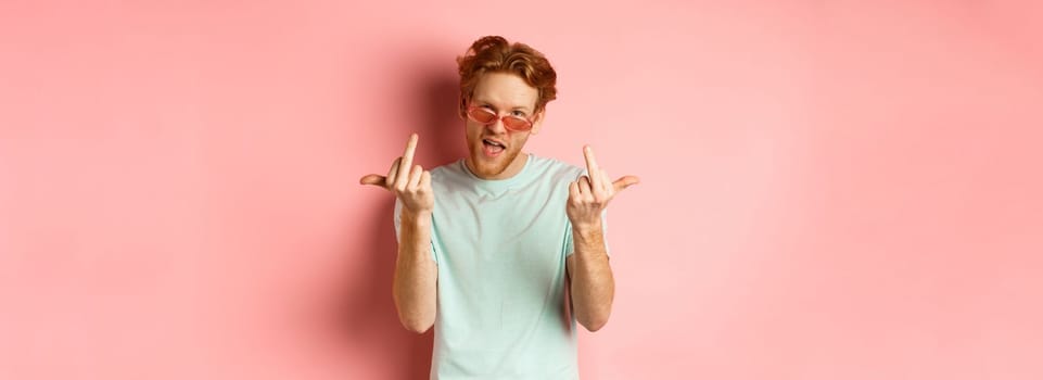 Arrogant and careless redhead man in sunglasses dont give a fuck, showing middle fingers at camera and frowning, standing over pink background.