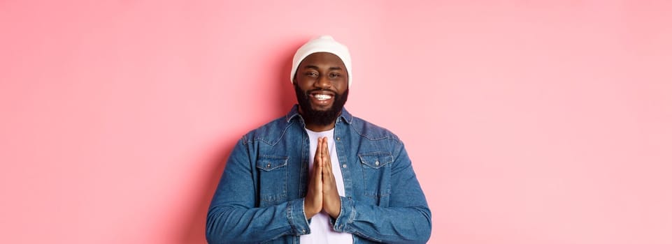 Happy smiling Black man saying thank you, holding hands in pray or namaste gesture, standing grateful against pink background.