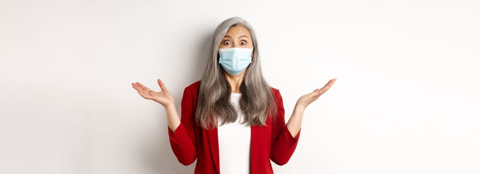 Covid-19 and business people concept. Asian businesswoman in face mask looking surprised, staring at camera in awe, standing over white background.