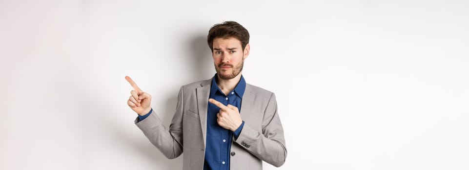 Showing bad example. Skeptical businessman look displeased and frowning while pointing at upper right corner banner, standing against white background.