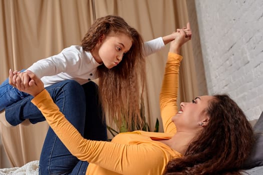 Love of young mother and daughter. They lie on the bed at home and have fun, they are fooling around, mother is holding her daughter on top of her legs