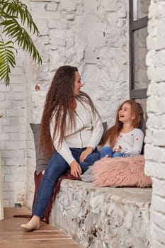 Mother and daughter sitting on sill near window in room. They show emotions and have fun