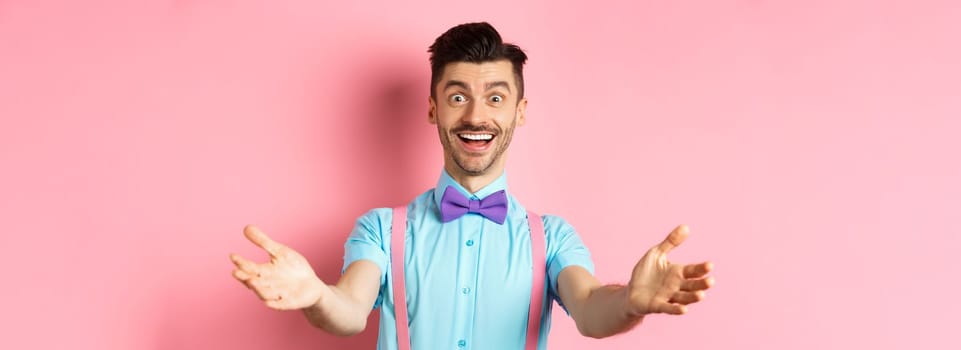 Happy handsome man in bow-tie stretch out hands in warm welcome, inviting or greeting you, receiving gift, standing on pink background.