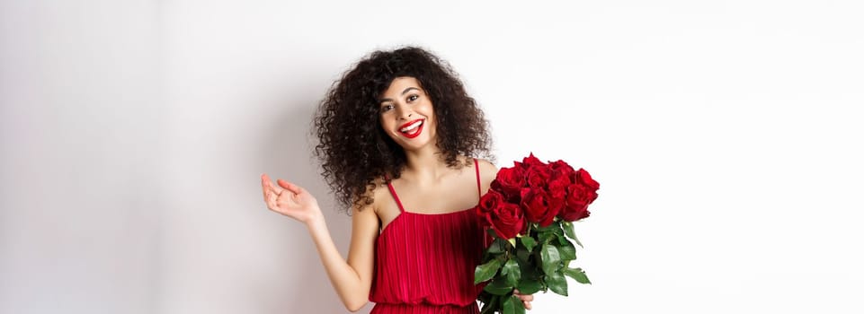 Happy woman celebrating, wearing stylish dress and holding flowers, smiling at camera, white background.