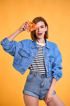 beautiful happy woman covering her eyes with a slice of fresh pizza on yellow background.