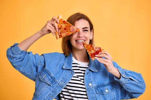 beautiful happy woman covering her eyes with slice of fresh pizza on yellow background.
