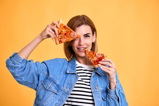 beautiful happy woman covering her eyes with slice of fresh pizza on yellow background.