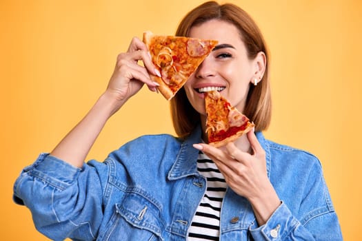 beautiful happy woman covering her eyes with slice of fresh pizza on yellow background.