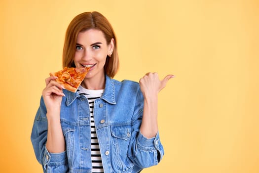 young woman with toothy smile eating delicious pizza slice and pointing at empty copy space on yellow background
