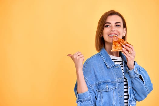 young woman eating delicious pizza slice and pointing at empty copy space on yellow background