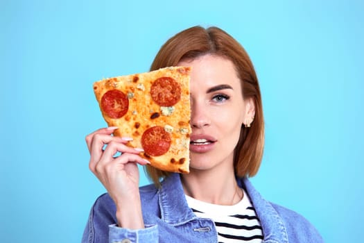 beautiful happy woman covering her eyes with a slice of fresh pizza on blue background.