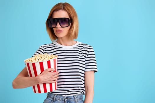 woman in sunglasses watching movie film, holding bucket of popcorn isolated on blue background. copy space