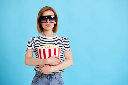 young woman in sunglasses watching movie film, holding bucket of popcorn isolated on blue background. copy space