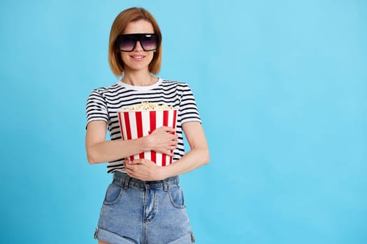 smiling woman in sunglasses and striped t-shirt jeans denim watching movie film, holding bucket of popcorn isolated on blue background. copy space