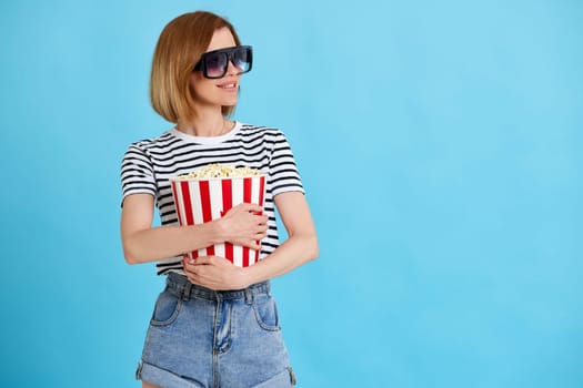 Cheerful young woman watching movie film, holding bucket of popcorn isolated on blue background. copy space