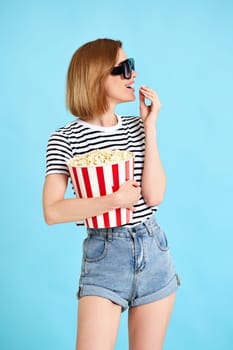 jouful caucasian woman in sunglasses watching movie film, eating popcorn isolated on blue background