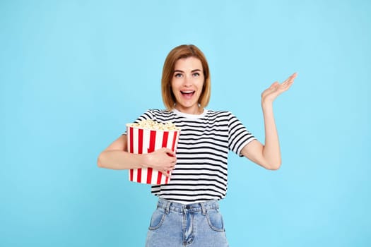 smiling beautiful woman in striped t-shirt jeans denim watching film, holding bucket of popcorn isolated on blue background