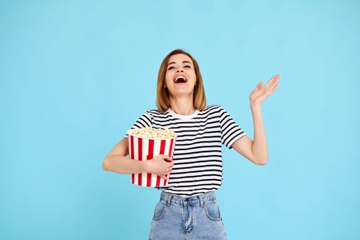 jouful caucasian woman watching movie film, holding bucket of popcorn isolated on blue background
