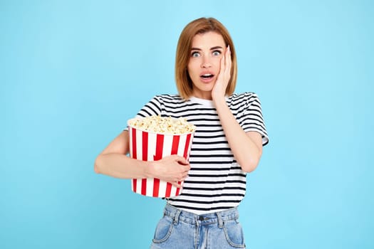 scared terrified woman watching movie film, holding bucket of popcorn isolated on blue background